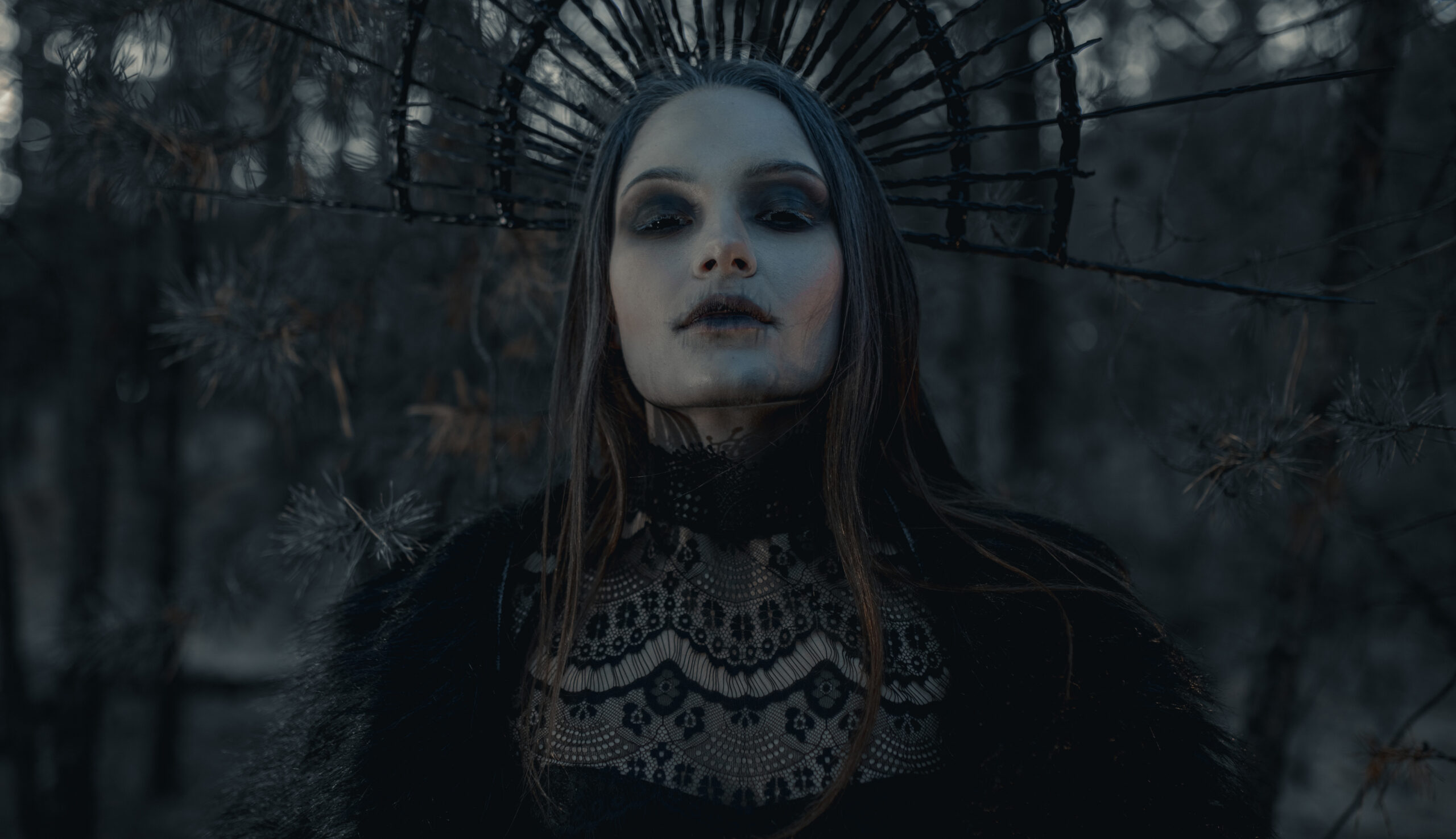 Portrait of young woman in image of witch in black dress and crown on her head in dark forest.